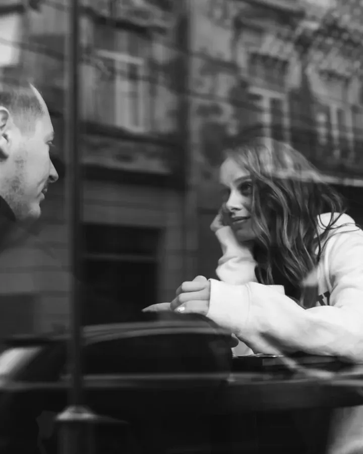 Un homme regarde une femme adossée à une voiture, dans une rue de ville, en noir et blanc